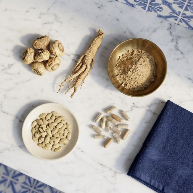 Raízes de maca e de ginseng, pó de raiz numa taça de latão, pevides de abóbora e cápsulas sobre mármore